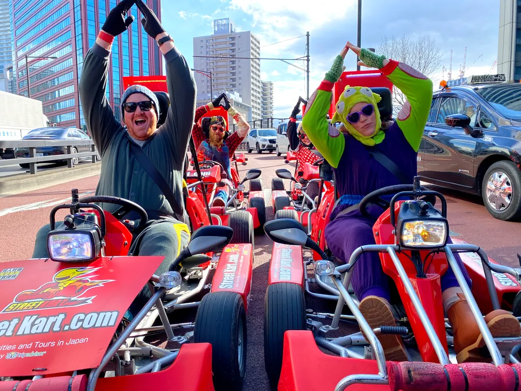 People in red go-karts on a city street, two in the foreground raising their arms in a 'Y' shape; one wears a frog hat and purple outfit.