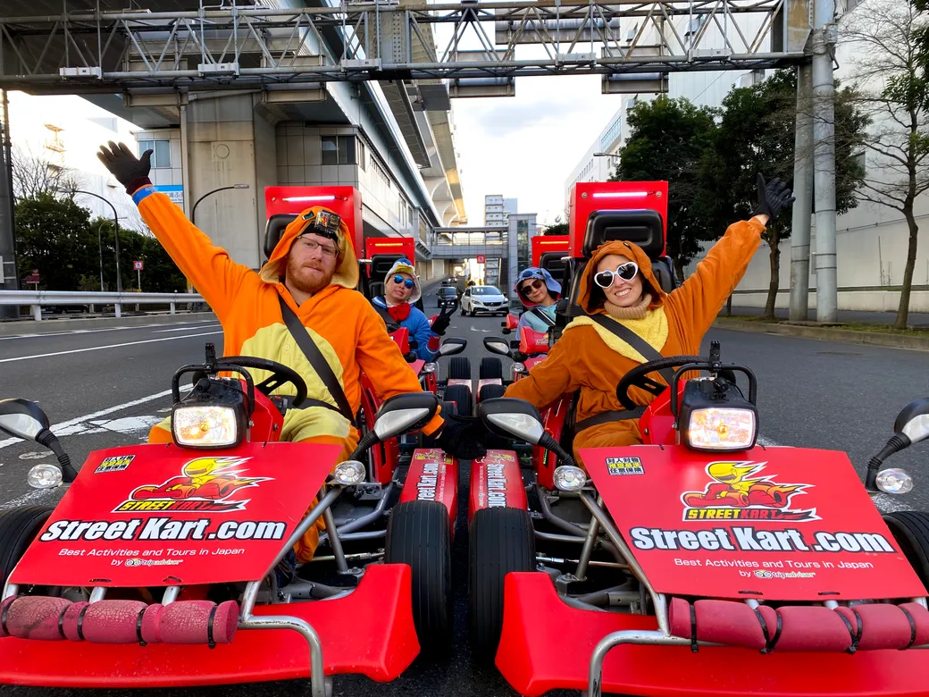 Two people in orange costumes drive bright red go-karts on a city street, arms raised, with StreetKart.com branding visible on the carts.