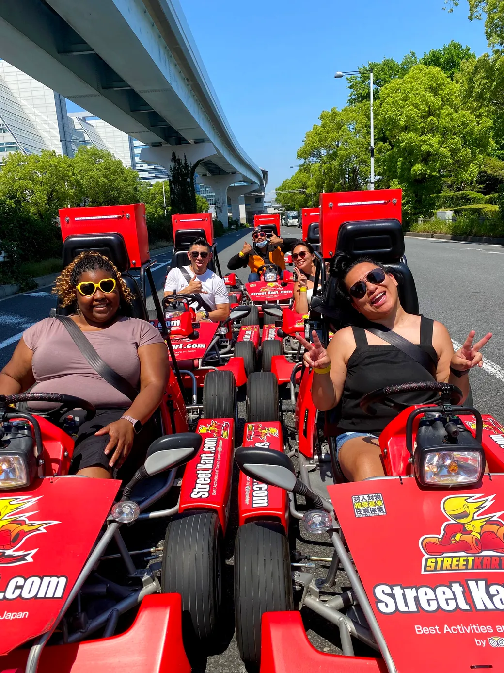 Six people in red go-karts on a sunny city street, posing under an elevated track above them.