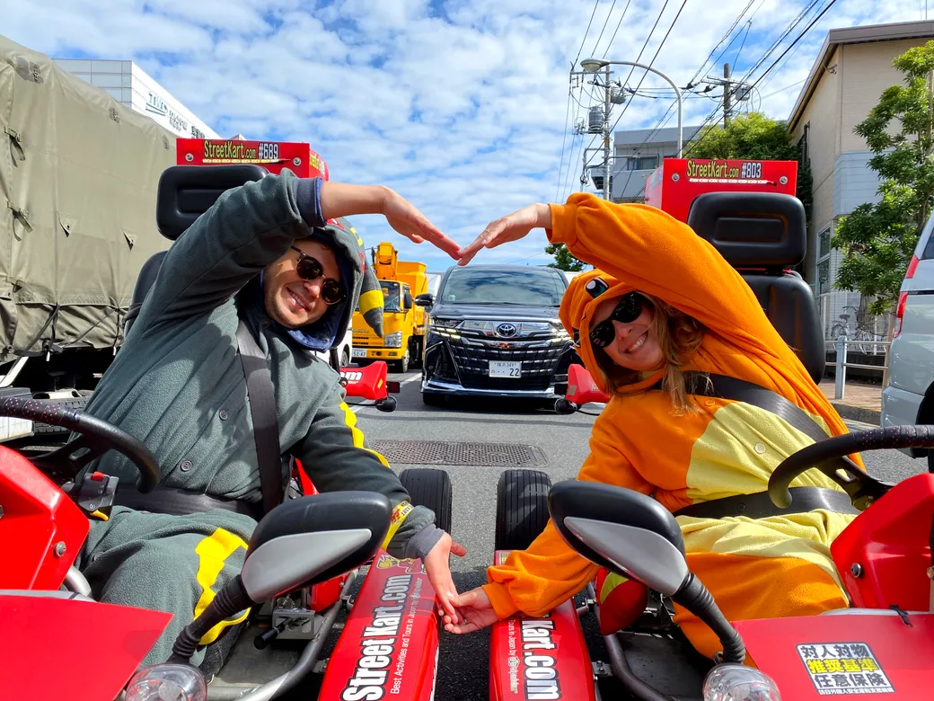 Two people in go-karts wearing colorful costumes lean toward each other and form a heart with their arms on a sunny street.